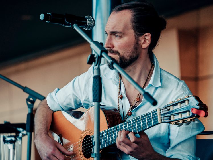 Image of A man with long hair tied back plays an acoustic guitar and sits behind a microphone on stage, wearing a light blue shirt and beaded necklace, looking thoughtfully to the side.