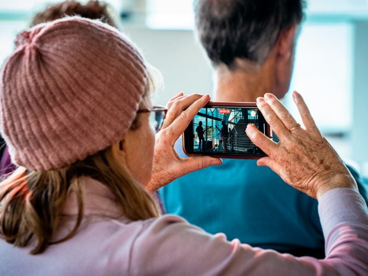 Image of A person wearing a pink knitted hat and glasses holds up a mobile phone, taking a photo or video of a scene through a window. The person is sitting among others, with the phone display showing a building and people walking.