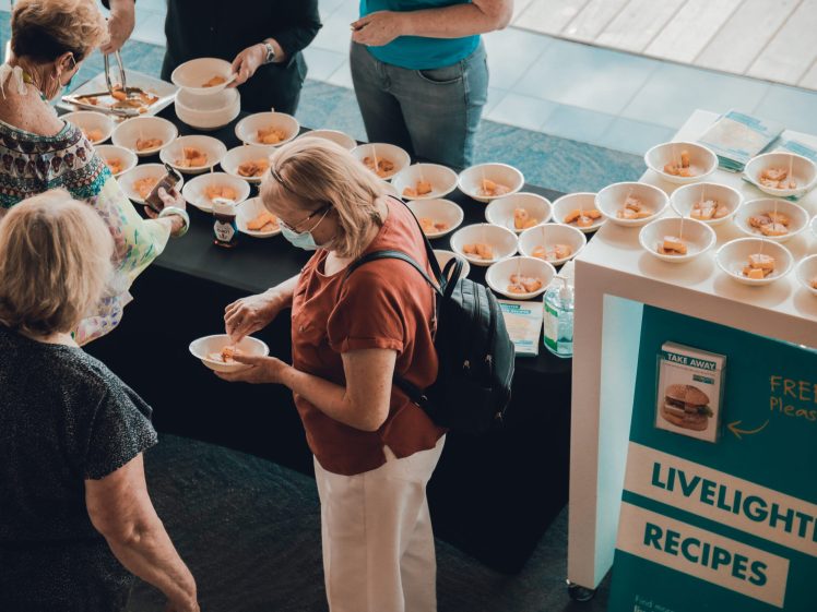 Image of People stand around a table with many bowls of food samples. A sign reads LiveLighter Recipes and offers free recipe cards. Some individuals are serving themselves, and the setting appears to be an indoor event or fair.
