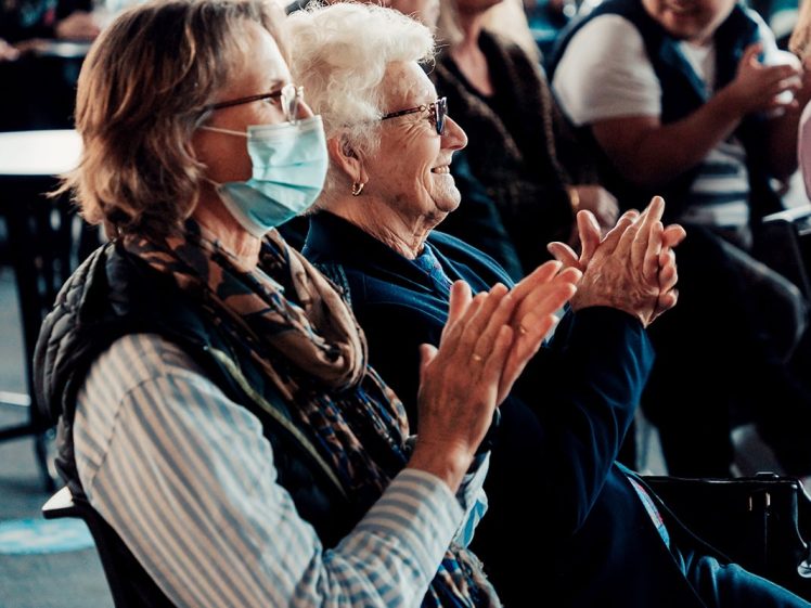 Image of Two older women sit side by side clapping at an event; one wears a scarf and a surgical mask, the other has white hair and spectacles. Other people are seated in the background, out of focus.