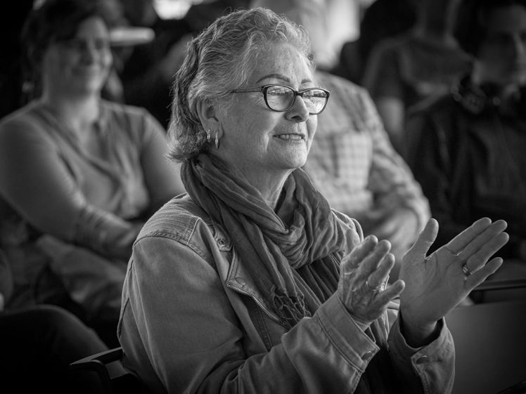Image of An older woman with glasses and a scarf smiles and claps whilst sitting in an audience. Other people are seated and blurred in the background. The photo is in black and white.