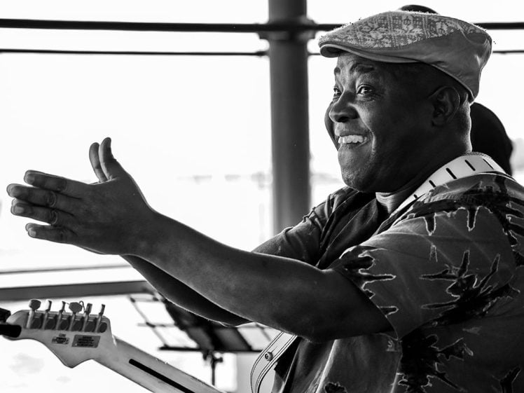 Image of A smiling man wearing a patterned shirt and flat cap claps his hands, holding an electric guitar, with bright light streaming in behind him. The photo is in black and white.