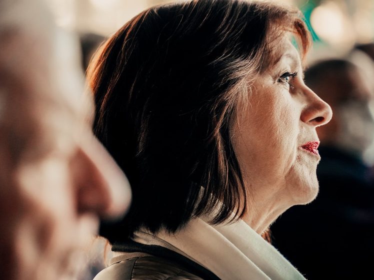 Image of A middle-aged woman with brown hair and a white scarf is shown in profile, looking ahead with a serious expression. She is surrounded by a blurred crowd, with a man partially visible in the foreground.