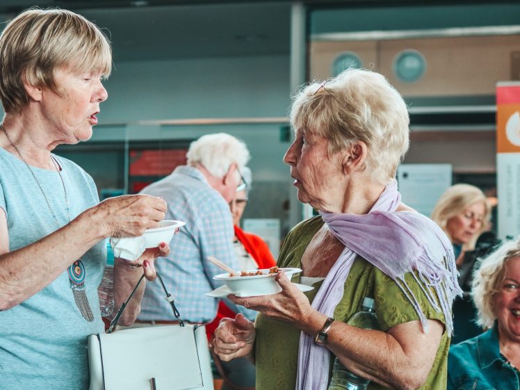 Image of Two older women stand and chat whilst eating at a social gathering. One woman gestures with her spoon, whilst the other holds a plate and water bottle. People are seated and smiling in the background.