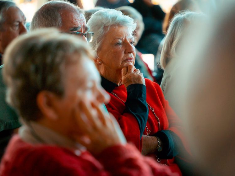 Image of A group of older adults sits closely together, listening attentively. The woman in the centre, wearing a red jumper, rests her chin on her hand, appearing thoughtful. The background is softly blurred.