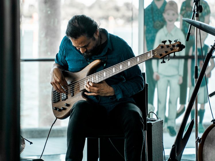 Image of A musician with dark hair and a beard sits on a chair, intently playing a six-string electric bass guitar. He wears a blue shirt and is indoors, with blurred people and outdoor scenery visible through large windows behind him.