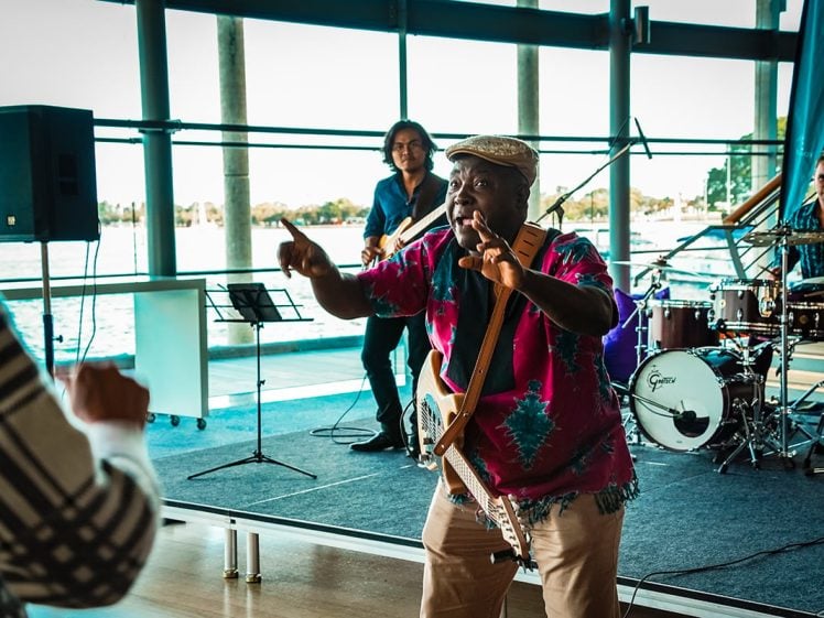 Image of A musician in a colourful shirt plays guitar and sings energetically to an audience indoors, with a band performing behind him near large windows overlooking water.