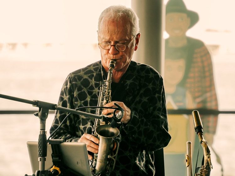 Image of An older man with glasses plays a saxophone indoors, seated behind a microphone and music stand. In the background, there is a standee of two cartoon figures.