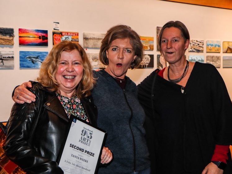 Image of Three women stand together smiling and looking surprised; one holds a Second Prize art award. They are in front of a wall displaying small colourful artworks.