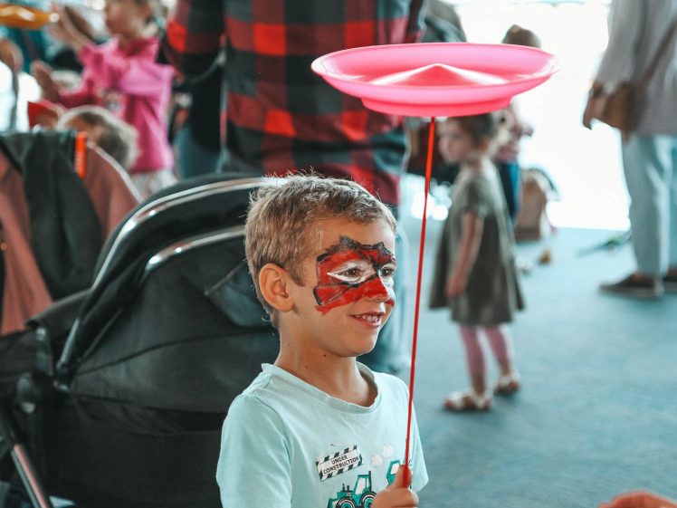 Image of A young boy with his face painted smiles whilst balancing a red spinning plate on a stick indoors, surrounded by other children and adults in the background.