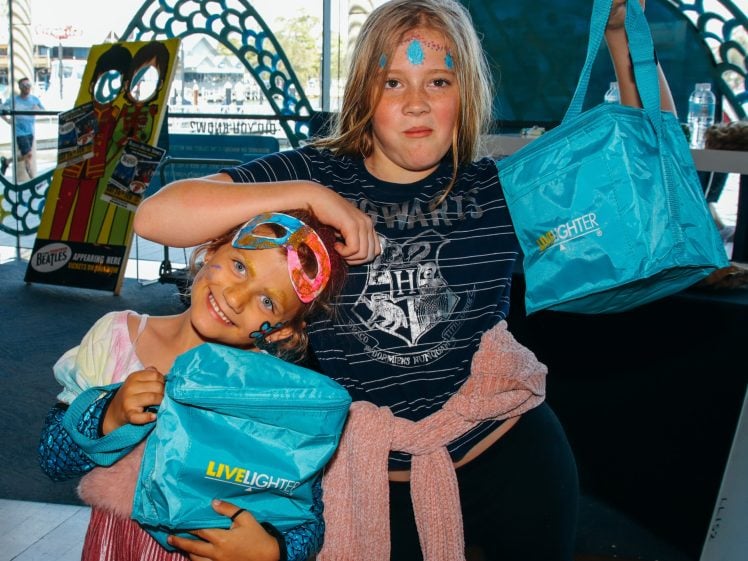 Image of Two children smiling and posing indoors, holding blue LiveLighter bags. One child wears a pink dress and colourful mask, the other a dark T-shirt with face paint. Bright light comes through the window behind them.