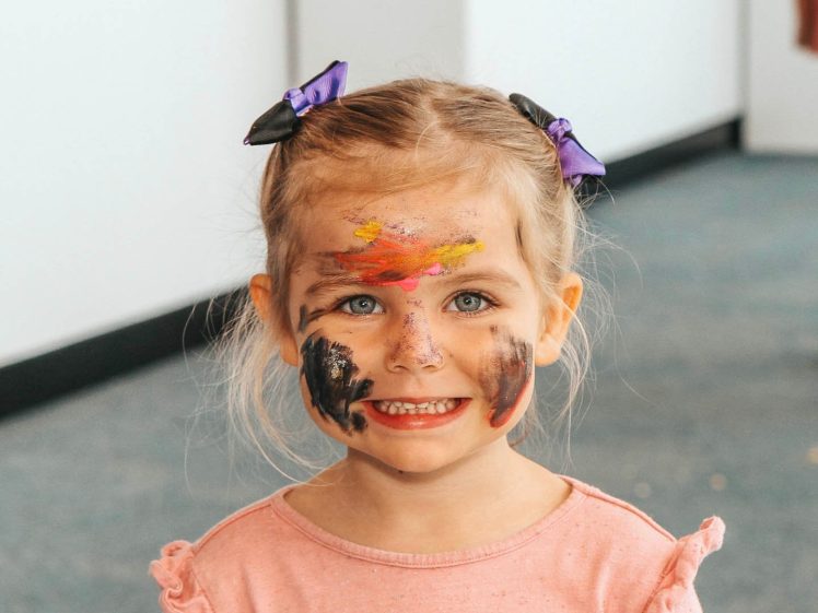 Image of A young girl with blue eyes and blonde hair in plaits with purple bows smiles. Her face is covered in colourful, messy face paint. She is wearing a light pink top and standing indoors.