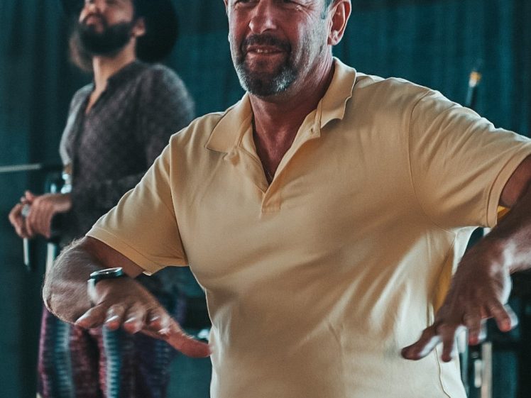 Image of A man in a yellow polo shirt dances with his hands raised, while another man with a beard and hat stands in the background on a stage with musical equipment.