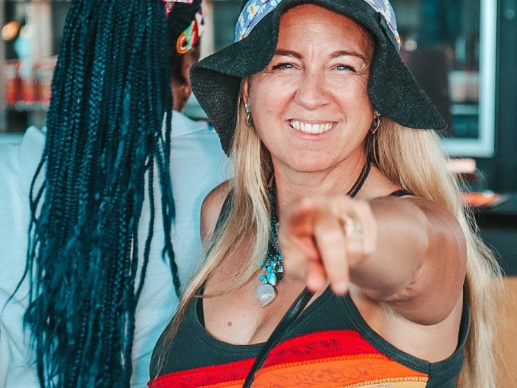 Image of A smiling woman with long blonde hair, wearing a rainbow-striped top and a floral bucket hat, points towards the camera. Another person with long plaited hair and a colourful outfit stands in the background, facing away.