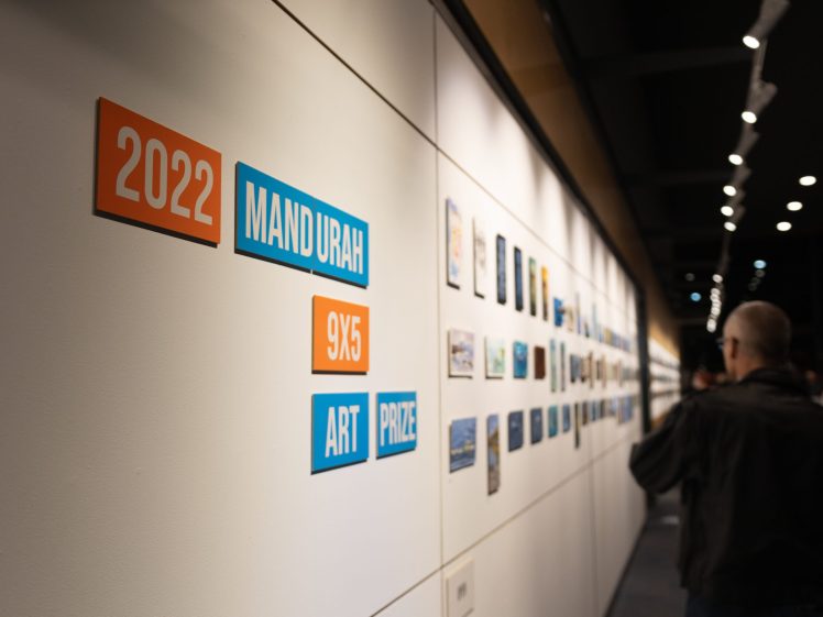 Image of A gallery wall displays small artworks with signs reading 2022 Mandurah 9x5 Art Prize on the left; several people are viewing the exhibits under spotlights.