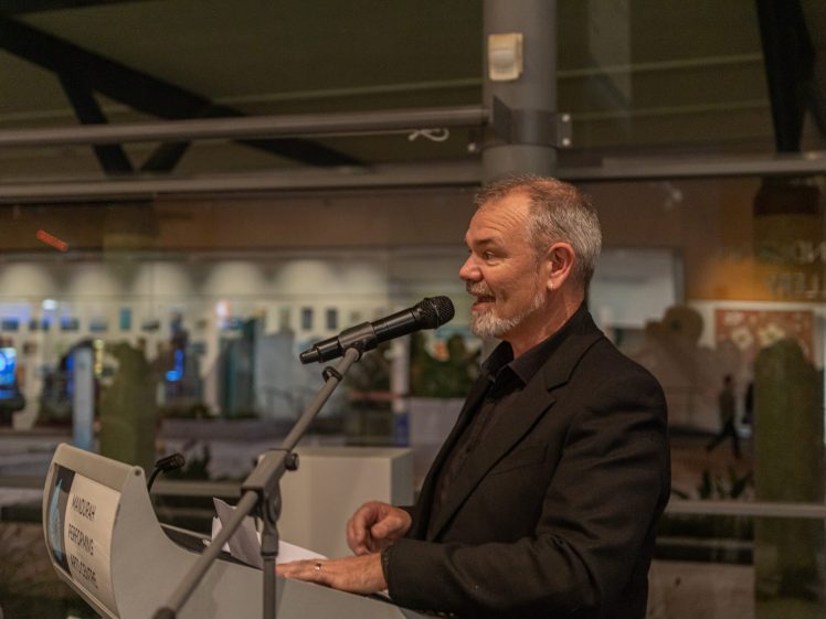 Image of A man in a black suit speaks into a microphone at a lectern during an indoor event. The background shows glass windows and some blurred lights.