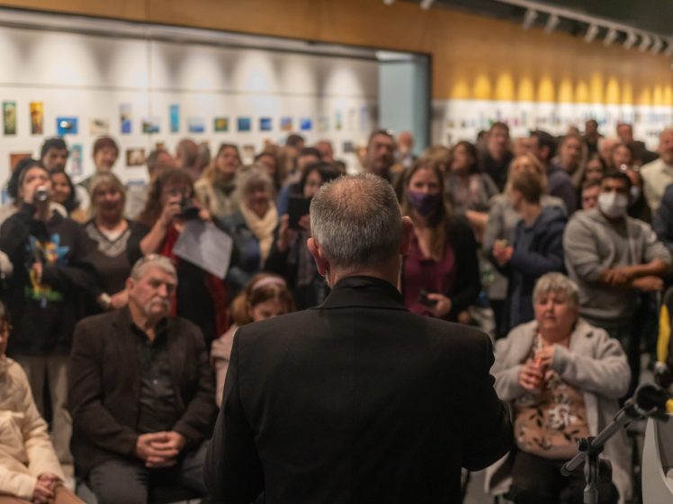 Image of A man in a suit speaks to a large, attentive crowd at an art gallery, with many people standing and viewing small artworks displayed on the walls behind them.