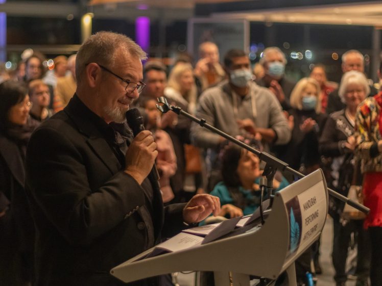 Image of A man speaks into a microphone at a lectern labelled VOIX DES USAGERS, addressing a large audience. People in the crowd watch attentively, with some wearing masks. The event is held indoors, with soft lighting.