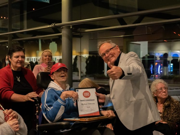 Image of A smiling man in a suit gives a thumbs-up while posing with a man in a wheelchair holding a framed certificate, surrounded by people, indoors at a public event.
