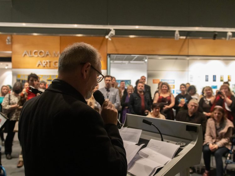 Image of A man speaks into a microphone at a lectern, facing a large audience in an art gallery. People in the crowd listen attentively, some holding phones and cameras. Artworks and signage are visible in the background.