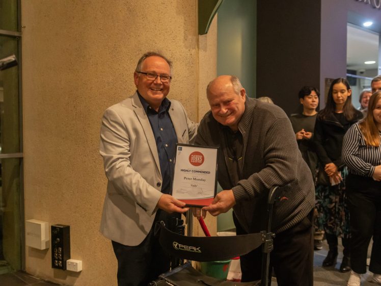Image of Two men smiling indoors, one in a light jacket handing a framed certificate to the other, who is leaning on a Zimmer frame. Several people stand in the background watching the presentation.