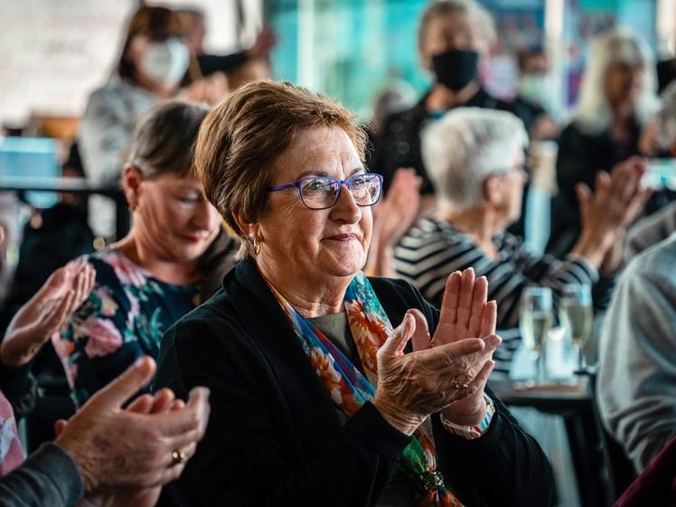 Image of A group of older adults sit closely together, clapping and smiling at an indoor event. The focus is on a woman with glasses and a colourful scarf, while others talk or watch in the background. Some people wear masks.