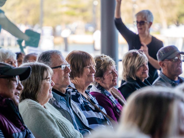 Image of A group of older adults sit closely together in an outdoor setting, smiling and listening attentively. One woman in the background is standing and raising her hand, and the atmosphere appears lively and engaged.