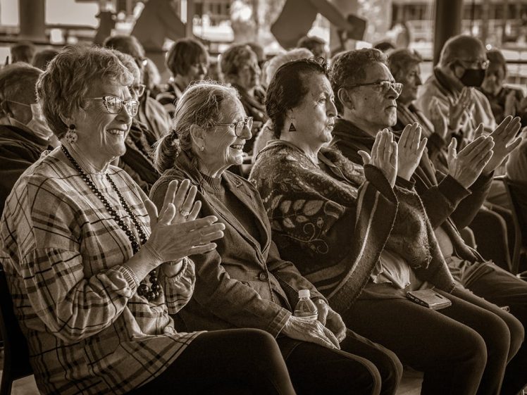 Image of A group of older adults sit in rows, smiling and clapping as they watch an event. The image is in sepia tone, emphasising the joyful, engaged expressions of the audience.