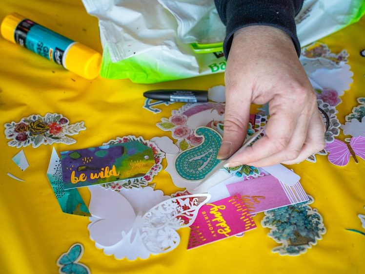 Image of A person’s hand arranges colourful decorative paper cut-outs, stickers, and cards with phrases like “be wild” on a yellow surface. A glue stick and scattered art supplies are also visible.