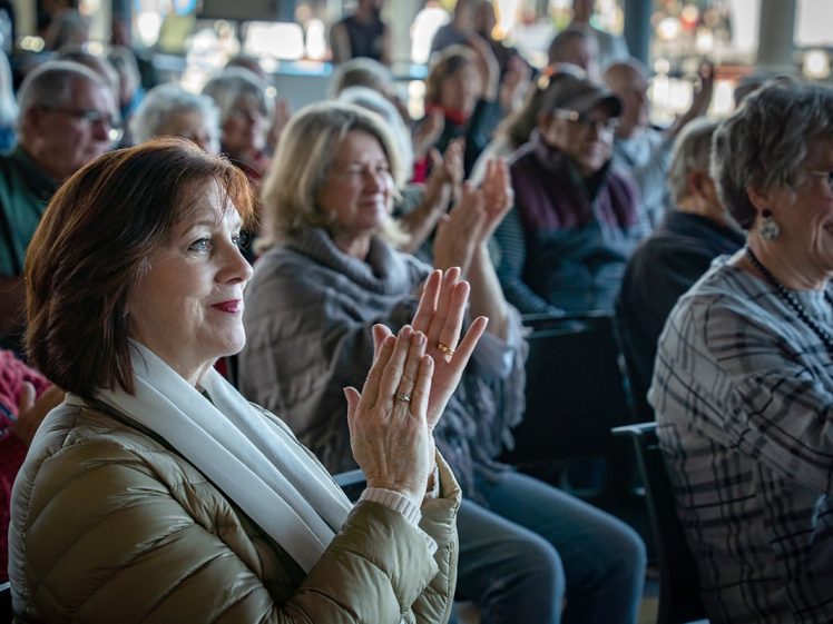 Image of A group of older adults sit closely together indoors, attentively watching something off-camera. Many are smiling and clapping, dressed in warm clothing, suggesting a positive and engaging event.
