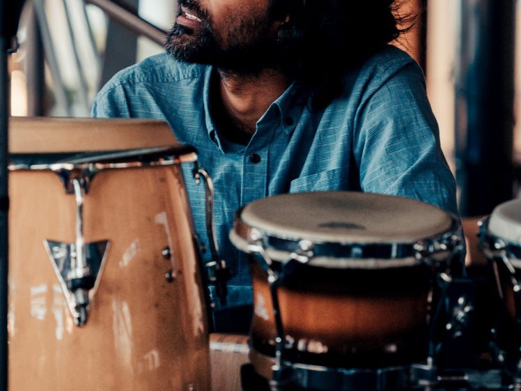 Image of A man with shoulder-length dark hair and a beard sits behind a set of conga and bongo drums, looking thoughtfully to his left. He is wearing a blue button-up shirt in a warmly lit indoor setting.