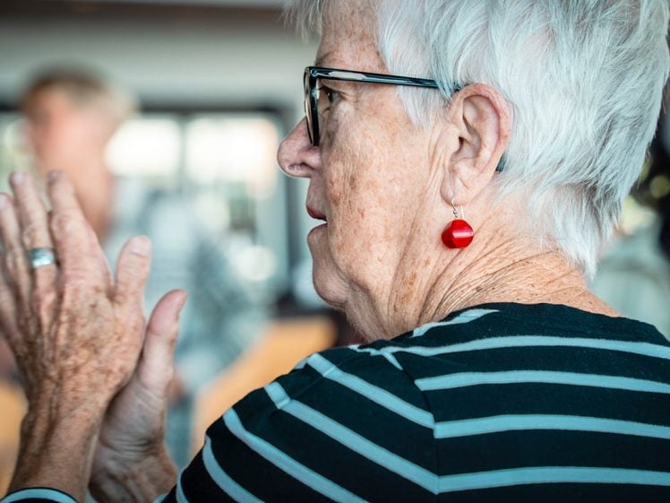 Image of An elderly woman with short white hair and glasses, wearing a black-and-white striped shirt and red earrings, is clapping her hands. The background is blurred.