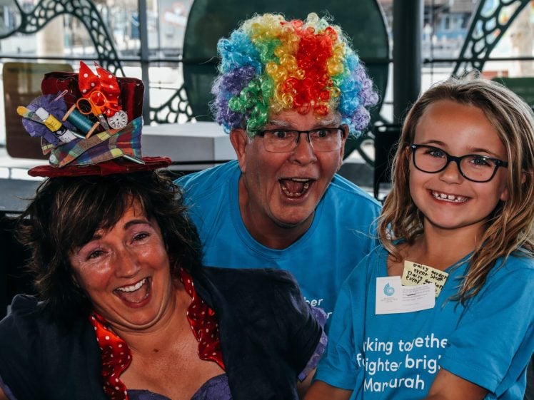 Image of Three people smiling and posing together; one woman wears a whimsical hat, a man is in a rainbow clown wig, and a girl with glasses stands beside them. All three wear blue shirts and appear to be at a festive event.