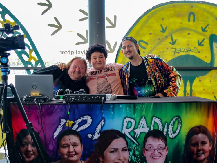 Image of Three people sit together at a colourful LIPA Radio table, smiling at the camera. Behind them, bright graffiti-style art decorates the windows, adding a lively backdrop to the radio set-up.