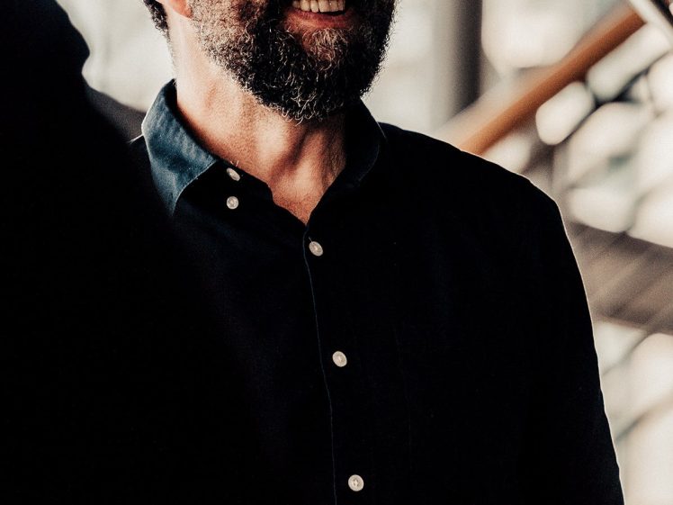 Image of A bearded man in a dark button-up shirt smiles and squints his eyes whilst standing indoors under natural light, with a blurred background and a microphone visible above him.
