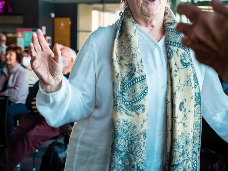 Image of An older woman with short grey hair and a patterned scarf smiles and raises her hands while dancing indoors, with people seated and watching in the background.