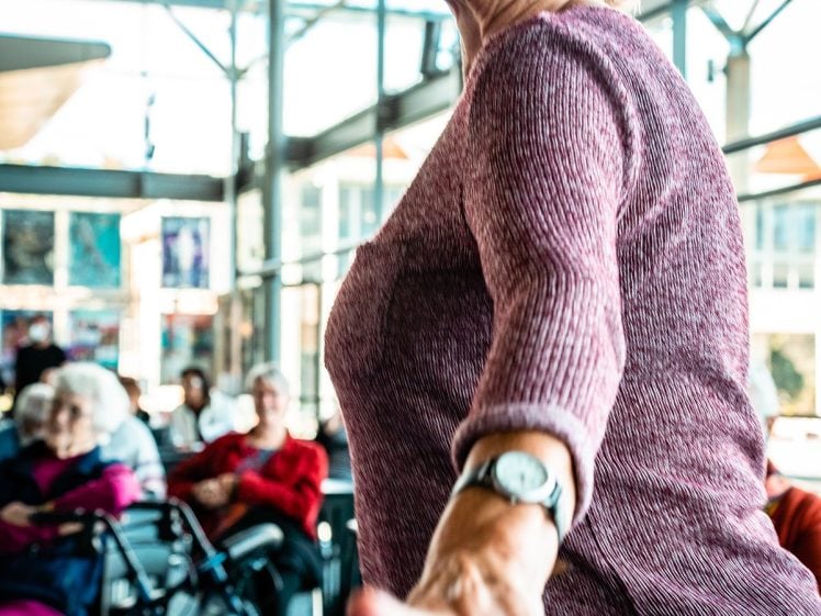 Image of An older woman in a purple top dances with a joyful expression in a bright room, while other pensioners sit and watch in the background. A zimmer frame is visible near the seated group.