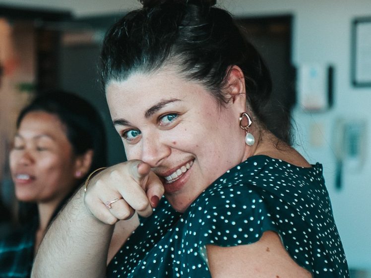 Image of A woman with dark hair in a bun, wearing a dark polka-dot top, smiles and points towards the camera. Another woman is in the background, slightly out of focus. They appear to be indoors, possibly at a social gathering.