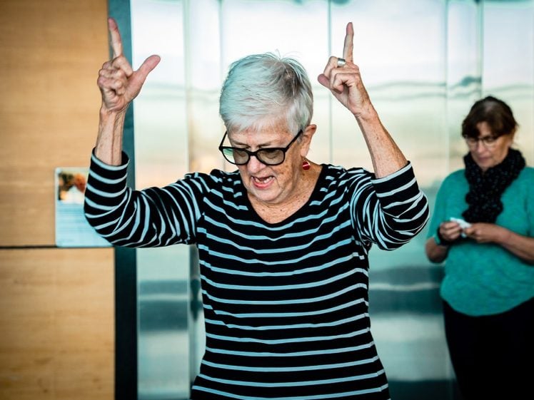 Image of An older woman with white hair and glasses, wearing a black-and-white striped top, raises both hands and points upwards. Another woman stands in the background, looking down at something in her hands.