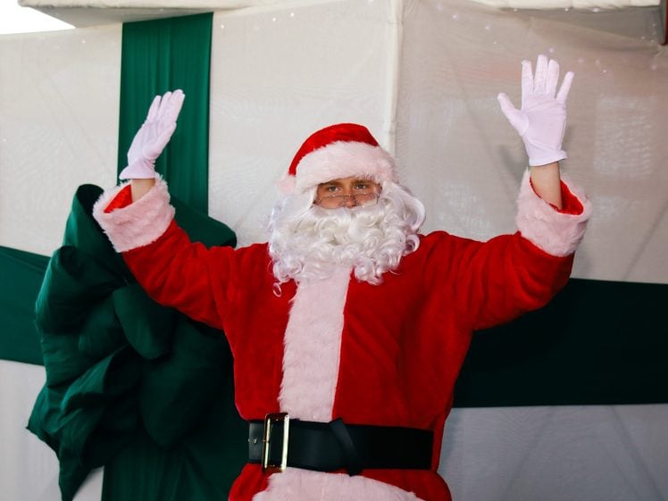 Image of A person dressed as Father Christmas with a white beard, red suit, and white gloves is raising both hands. Behind them is a large green ribbon decoration on a white background, resembling a wrapped present.