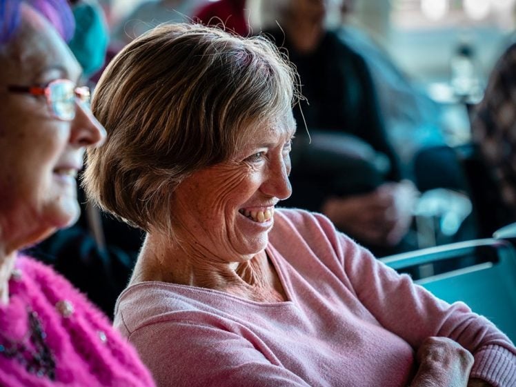 Image of Two older women sitting side by side, smiling and laughing whilst looking ahead. Both are wearing glasses and jumpers, with a group of people blurred in the background. The atmosphere appears friendly and relaxed.