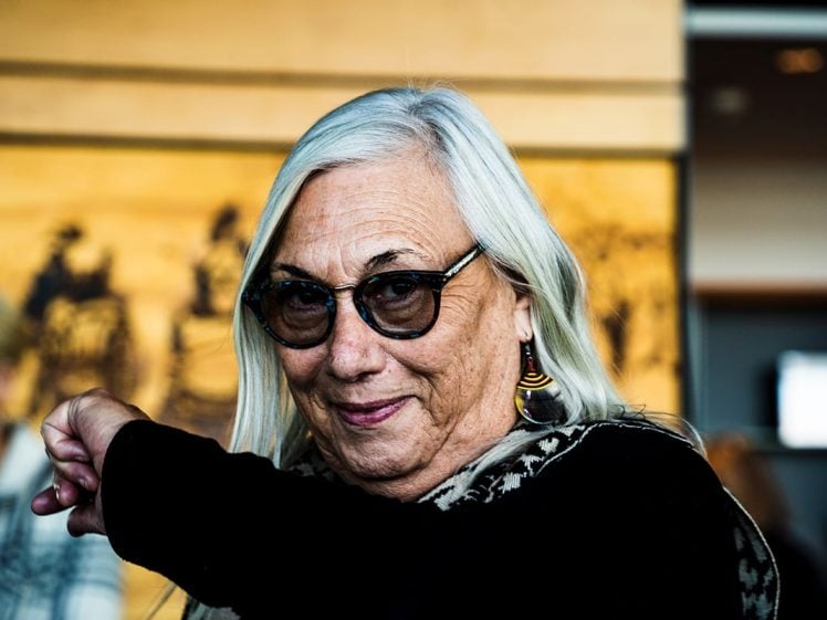 Image of An older woman with long grey hair and glasses smiles, wearing patterned earrings and a scarf. She is indoors in front of a yellow and brown background, with her arm raised in the foreground.