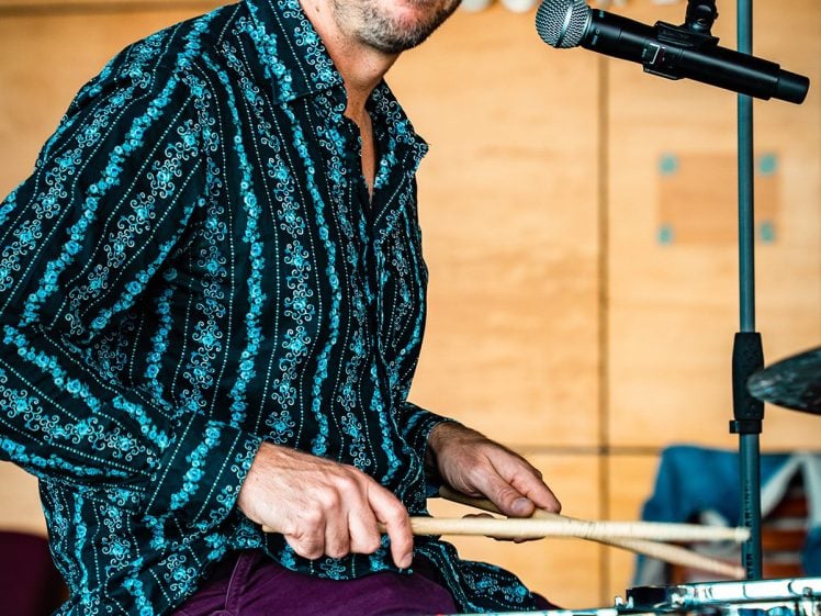 Image of A man in a patterned shirt plays the drums and looks at the camera, with a microphone positioned nearby and a wooden wall with numbers in the background.