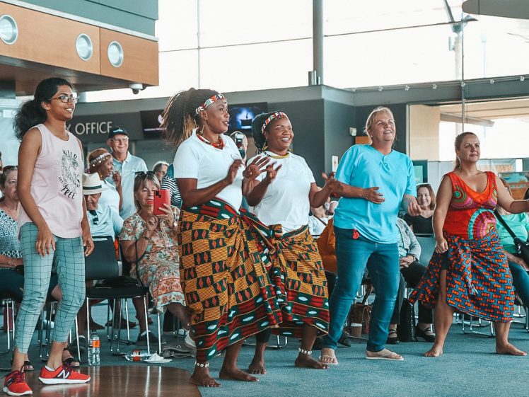Image of A group of five people stand and dance joyfully in front of an audience seated indoors. Three women wear colourful patterned skirts and headbands, while others are in casual clothing. Onlookers watch and smile in the background.