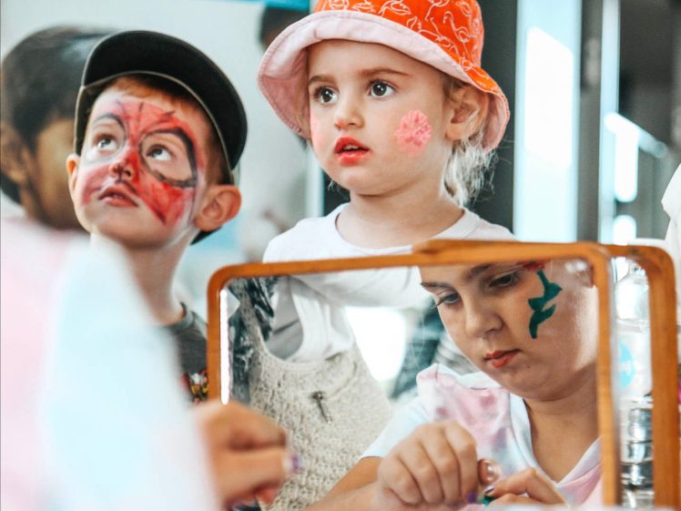 Image of Two young children with painted faces, one wearing an orange hat, look at themselves in a mirror. Another child paints their face in the reflection. All appear focused and engaged in the activity.