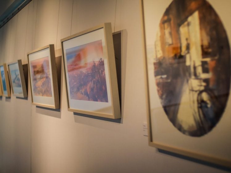 Image of Framed abstract paintings hang in a row on a gallery wall, slightly angled, with soft natural light coming from the left side of the image.