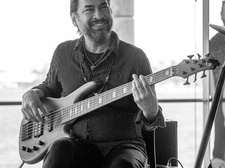 Image of A man with a beard and long hair smiles whilst playing an electric bass guitar, seated indoors near large windows with waterfront views. Microphone stands and audio equipment are visible nearby. Black and white photo.