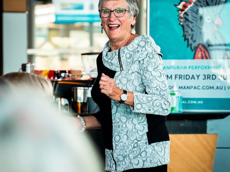 Image of An older woman with short grey hair, glasses, and a patterned top smiles and stands in a bright indoor setting, with a colourful poster and people in the background.