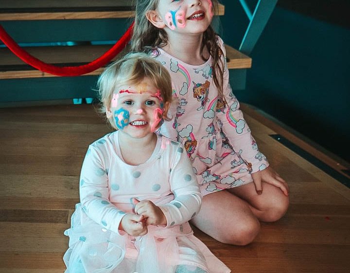 Image of Two young children with face paint smile while sitting on wooden stairs. The child in front wears a white dress with polka-dot leggings, and the child behind wears a pink dress with unicorns. Both look happy and cheerful.