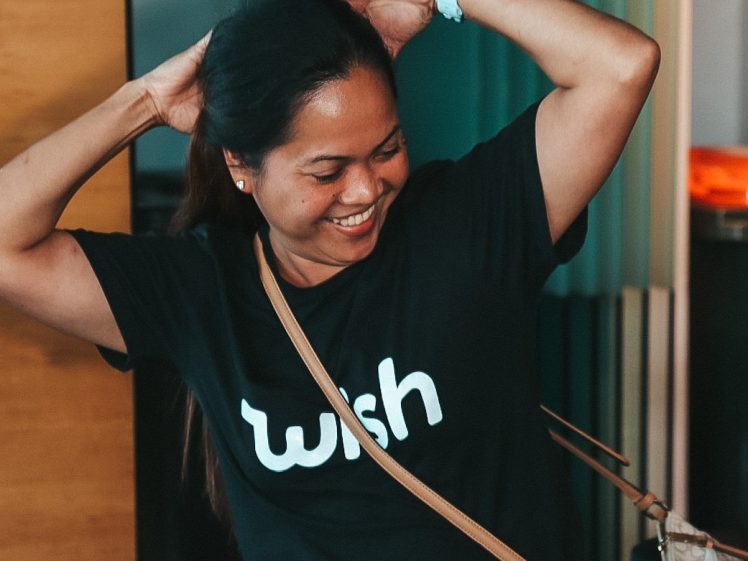 Image of A smiling woman wearing a black wish T-shirt poses playfully with her hands behind her head. She has long dark hair, blue jeans, and a tan handbag slung across her body.